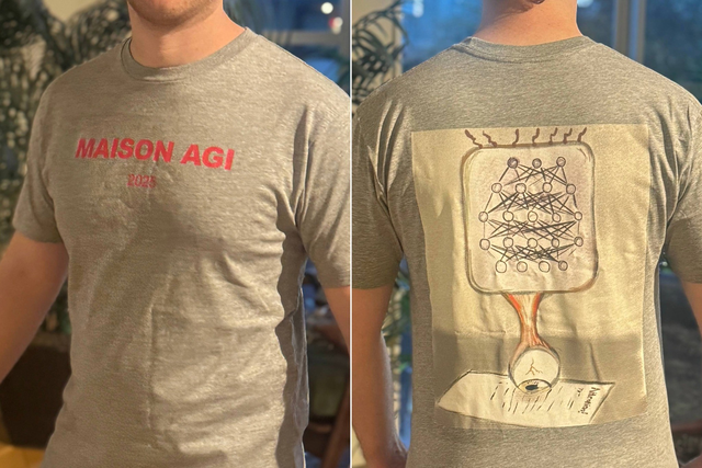 A person wears a gray t-shirt with “MAISON AGI 2023" in red on the front and a neural network diagram connected to an eyeball on paper on the back.